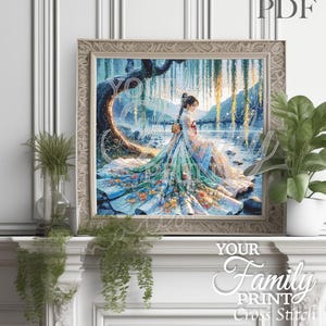 May include: A framed cross-stitch art print depicts a woman in a flowing gown seated near a body of water. The artwork features a detailed design with blue, green, and floral accents. The frame is a light beige color. The text "Your Family Print Cross Stitch" is visible.