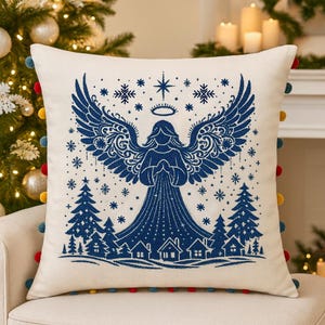 Puede incluir: Cojín decorativo crema con un diseño de ángel azul, alas y una escena de pueblo. El cojín tiene un ribete de pompón colorido. Es un artículo de decoración navideña festivo.