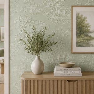 May include: A light green wallpaper with a white embossed toile pattern featuring deer and trees. A white ceramic vase holds green branches next to a stack of three books titled Live Beautifully, This Is Home, and The Kinfolk Home with a small bowl.