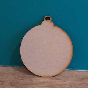 Puede incluir: Un adorno redondo de madera liso, con un pequeño agujero en la parte superior para colgar. El adorno es de color marrón claro y tiene una superficie lisa.