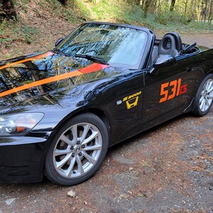 以下が含まれることがあります： 黄色のオレンジ色のレーシングストライプがフードに施された黒いホンダS2000コンバーチブル。車には側面に「S2000」のエンブレムとドアに「531」の番号が付けられています。