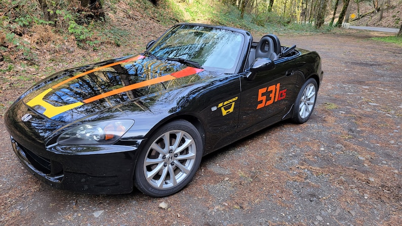 以下が含まれることがあります： 黄色のオレンジ色のレーシングストライプがフードに施された黒いホンダS2000コンバーチブル。車には側面に「S2000」のエンブレムとドアに「531」の番号が付けられています。
