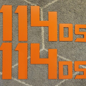 以下が含まれることがあります： オレンジ色の数字と文字「1140s」が2セットあり、質感のある素材でできています。数字と文字は水平に並んでいます。