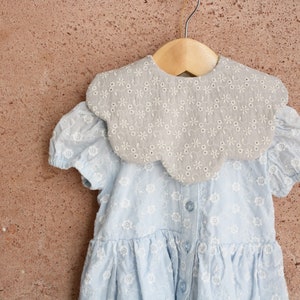 K&ouml;nnte beinhalten: Ein hellblaues Kleid mit einem gekr&auml;uselten, bestickten Kragen. Das Kleid hat ein Blumenmuster und h&auml;ngt an einem Holzb&uuml;gel.