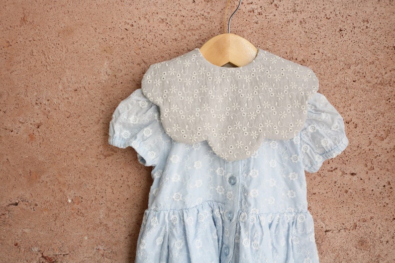 K&ouml;nnte beinhalten: Ein hellblaues Kleid mit einem gekr&auml;uselten, bestickten Kragen. Das Kleid hat ein Blumenmuster und h&auml;ngt an einem Holzb&uuml;gel.