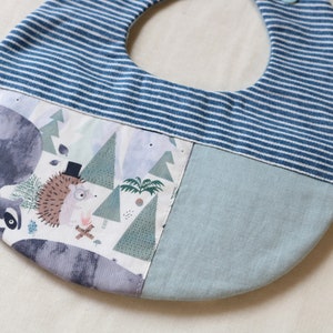 May include: A blue and white striped fabric baby bib with a woodland animal print. The bib features a raccoon, a hedgehog, and trees. The bib has a blue and white striped top section and a light blue bottom section. The bib has a small white tag with the word "Kizara" on it.