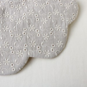 K&ouml;nnte beinhalten: Nahaufnahme eines grauen Stoffes mit wei&szlig;en gestickten Blumen. Der Stoff hat einen gewellten Rand.
