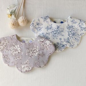 Könnte beinhalten: Zwei Lätzchen mit Blumenmuster. Eines ist lavendelfarben mit weißen Blumen, das andere weiß mit blauen Blumen. Beide haben einen gewellten Rand und sind aus Stoff. Die Lätzchen sind für Babys.