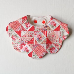 May include: A patchwork baby bib with a scalloped edge. The bib features a variety of red and pink floral patterns and geometric shapes. Two red buttons secure the bib at the neck. The bib is designed for infants.