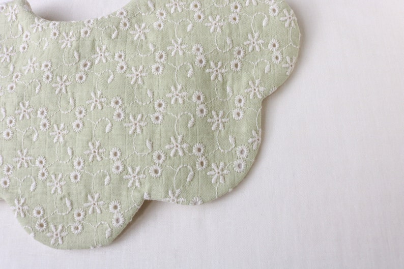 K&ouml;nnte beinhalten: Ein hellgr&uuml;ner Baby-L&auml;tzchen mit einem gewellten Rand und einem wei&szlig;en, gestickten Blumenmuster.