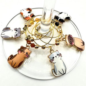 May include: Set of six gold-toned wine glass charms featuring different cartoon cats. Each charm has a different colored cat with a white background.