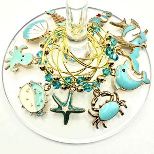 May include: Wine glass charms with a sea life theme. The charms feature a crab, starfish, dolphin, and fish, in turquoise, white, and gold. The charms are attached to gold-coloured rings.