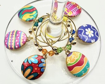 May include: Set of six colourful Easter egg-shaped wine glass charms. Each charm features a different patterned design with bright colours like pink, blue, green, yellow, and orange.