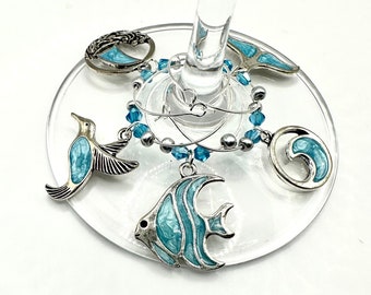 Könnte beinhalten: Satz mit fünf blauen und silbernen Weinglasanhängern mit einem Fisch, einer Welle, einem Kolibri, einem Seepferdchen und einem Baum. Die Anhänger sind an einer silbernen Kette mit blauen Perlen befestigt.