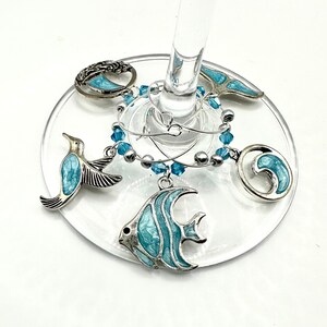 May include: Set of five blue and silver wine glass charms featuring a fish, a wave, a hummingbird, a seahorse, and a tree. The charms are attached to a silver chain with blue beads.