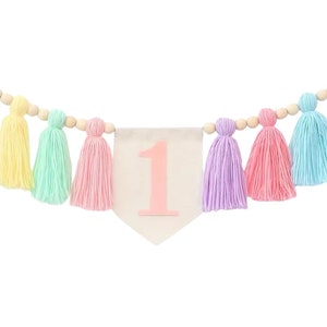 May include: A pastel-colored tassel garland with a white banner featuring the number "1" in pink. The garland is strung with wooden beads and features tassels in yellow, green, pink, purple, and blue.
