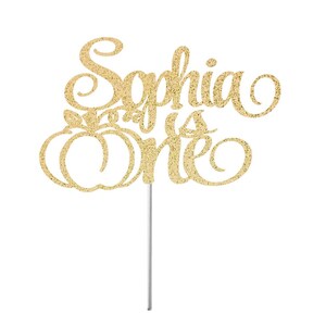 May include: Gold glitter cake topper with the text "Sophia is one" and a pumpkin design.