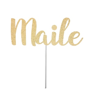 May include: Gold glitter cake topper with the word "Maile" in a cursive font.