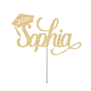 May include: A gold glitter cake topper with the name "Sophia" in cursive script. A graduation cap with the year "2026" is above the name. The topper is attached to a thin, silver stick.
