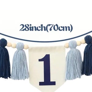 K&ouml;nnte beinhalten: Ein wei&szlig;es Banner mit der Zahl 1 in Blau, umgeben von einer Girlande aus Holzperlen und blauen und wei&szlig;en Garnquasten. Das Banner ist 70 cm lang.