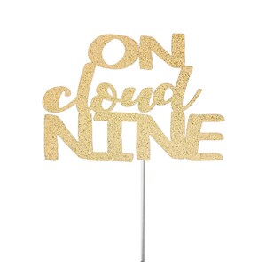 May include: A gold glitter cake topper with the phrase "ON cloud NINE" in a decorative font. The topper is attached to a thin, silver-colored stick, perfect for adding a festive touch to a cake or dessert.