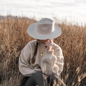 Op de afbeelding: Een persoon met een witte fedora-hoed en een beige trui zit in een veld met hoog, droog gras. De persoon draagt een gouden ketting met een ronde hanger.