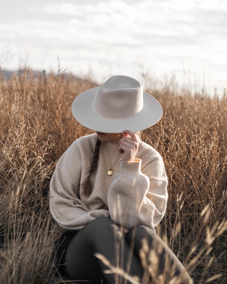 Op de afbeelding: Een persoon met een witte fedora-hoed en een beige trui zit in een veld met hoog, droog gras. De persoon draagt een gouden ketting met een ronde hanger.