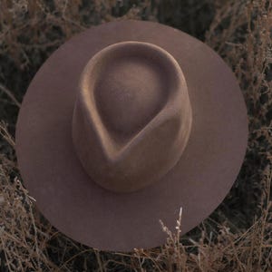 Puede incluir: Un sombrero fedora de ala ancha en un color malva apagado, sobre un fondo de hierbas beige secas. El sombrero presenta una corona estructurada y un ala ligeramente curvada, ofreciendo una silueta clásica. El color del sombrero se mezcla con el entorno natural.