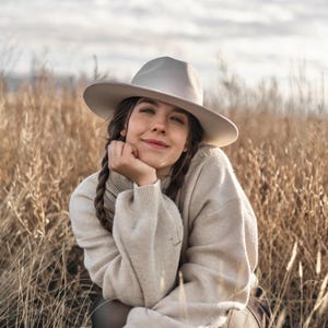 Op de afbeelding: Een vrouw in een lichtbeige trui en een lichtgrijze fedora zit in een veld met hoog, droog gras. Ze glimlacht en kijkt naar de camera.