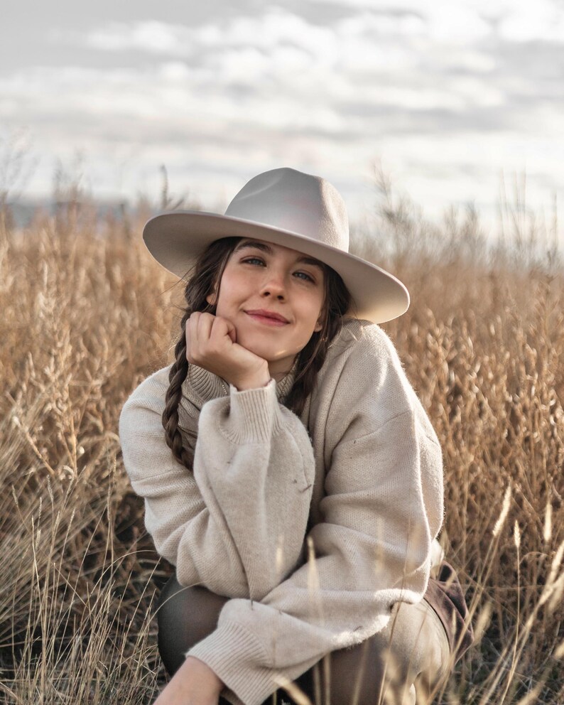 Op de afbeelding: Een vrouw in een lichtbeige trui en een lichtgrijze fedora zit in een veld met hoog, droog gras. Ze glimlacht en kijkt naar de camera.