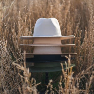 Op de afbeelding: Een stapel van vier hoeden in een veld met droog gras. De bovenste hoed is wit, de tweede hoed is beige, de derde hoed is zwart en de onderste hoed is groen.