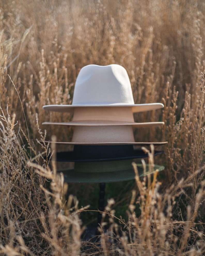 Op de afbeelding: Een stapel van vier hoeden in een veld met droog gras. De bovenste hoed is wit, de tweede hoed is beige, de derde hoed is zwart en de onderste hoed is groen.