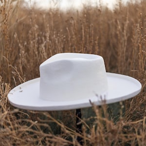 May include: A white felt fedora hat with a wide brim, sitting on a bed of dry grass.