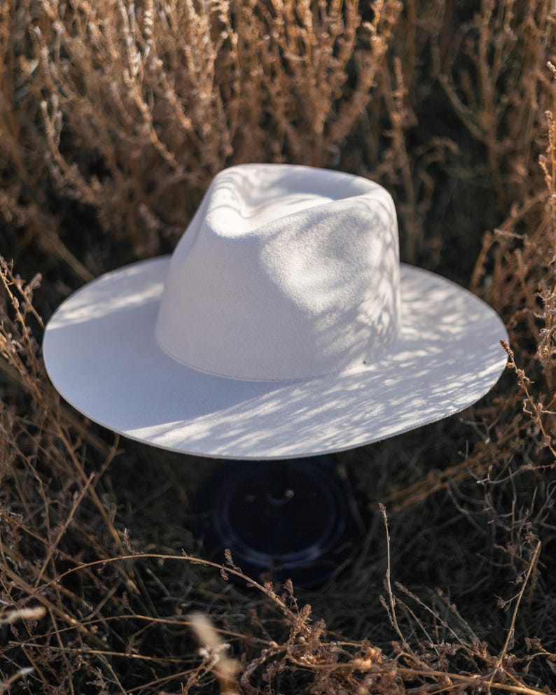 Op de afbeelding: Een witte fedora hoed met een brede rand, die op een donker oppervlak in een veld van droog gras ligt.
