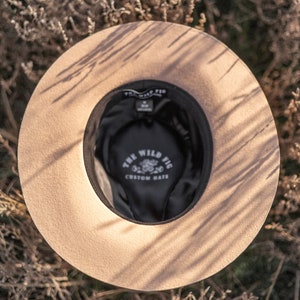 Op de afbeelding: Een beige wollen fedora hoed met een zwarte band en de tekst "The Wild Fig Custom Hats" in witte letters aan de binnenkant van de hoed.