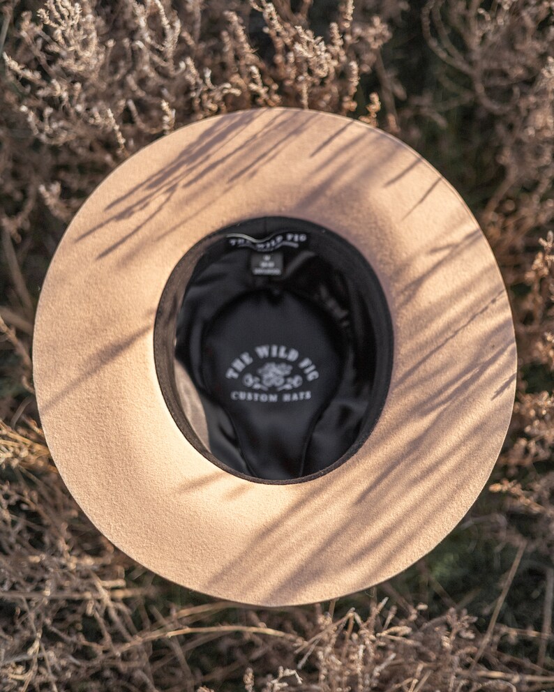 Op de afbeelding: Een beige wollen fedora hoed met een zwarte band en de tekst "The Wild Fig Custom Hats" in witte letters aan de binnenkant van de hoed.