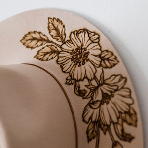 Op de afbeelding: Een beige hoed met een bloemmotief dat in de rand is gebrand. De bloemen zijn bruin en lijken op rozen.