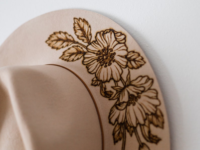 Op de afbeelding: Een beige hoed met een bloemmotief dat in de rand is gebrand. De bloemen zijn bruin en lijken op rozen.