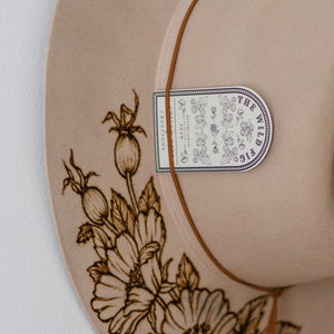 Op de afbeelding: Een beige wollen hoed met een bruin leren band en een bloemenpatroon. De hoed heeft een label met de tekst "The Wild Fig Creations".