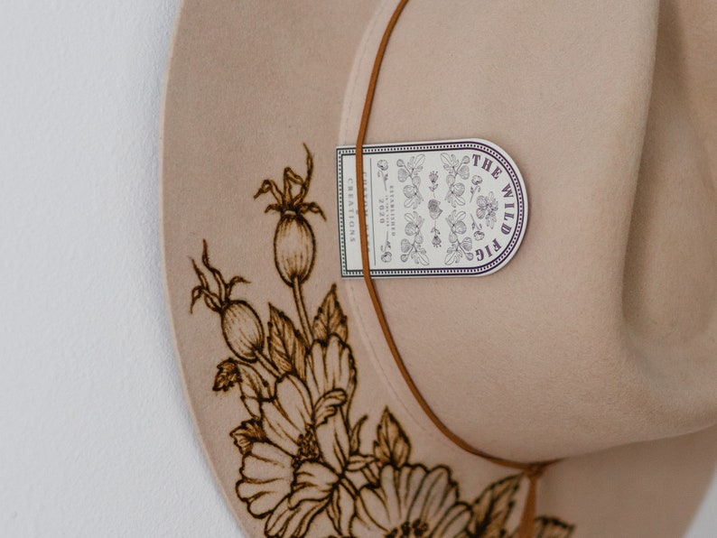 Op de afbeelding: Een beige wollen hoed met een bruin leren band en een bloemenpatroon. De hoed heeft een label met de tekst "The Wild Fig Creations".
