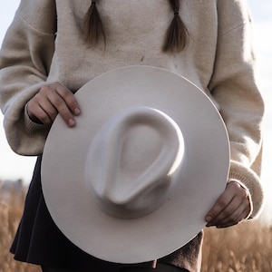 Puede incluir: Una persona con un suéter beige y una falda negra sostiene un gran sombrero fedora de fieltro beige claro.