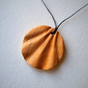 May include: A wooden pendant necklace with a round, textured, light brown wood pendant. The pendant has a small hole at the top for the black cord.