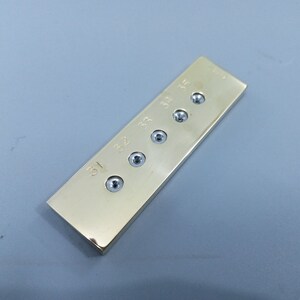 Può includere: Un oggetto rettangolare in metallo color oro con cinque teste di viti incassate. I numeri 31, 32, 33, 34 e 35 sono incisi sulla superficie. L'oggetto è posto su uno sfondo grigio.