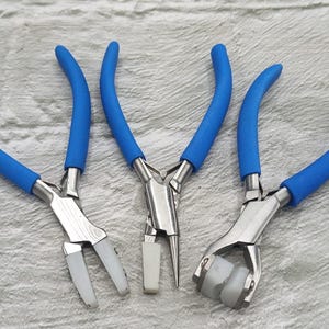 Könnte beinhalten: Drei Schmuckzangen mit blauen Griffen und silberfarbenen Metallkomponenten. Die Zangen haben verschiedene Kopfformen, flach, spitz und abgerundet, mit weißen oder grauen Spitzen. Werkzeuge für die Schmuckherstellung.