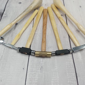 Könnte beinhalten: Eine Anordnung von Hämmern mit Holzgriffen und Metallköpfen, die in einem Halbkreis angeordnet sind. Die Werkzeuge weisen unterschiedliche Formen und Größen auf. Der Hintergrund ist eine helle Holzoberfläche, vermutlich für Schmuckarbeiten.