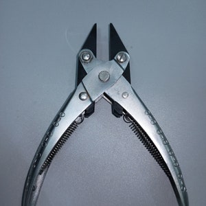 May include: Silver wire cutter pliers with black cutting edges. The handles are curved with small, circular indentations. A coiled spring is visible between the handles. The tool is designed for cutting wire.