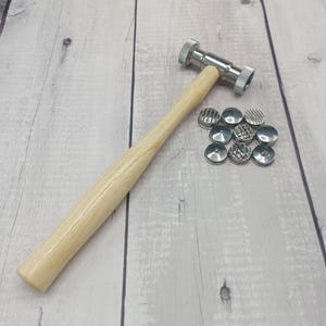 Può includere: Un martello in metallo con manico in legno chiaro e un set di nove timbri in metallo strutturato. Il martello ha una testa argentata e un manico in legno chiaro. I timbri hanno vari disegni.