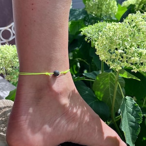 Può includere: Una cavigliera giallo neon con una piccola perla scura. La cavigliera è indossata su una caviglia, con una superficie in legno e fogliame verde sullo sfondo. La cavigliera ha un design semplice e minimalista.