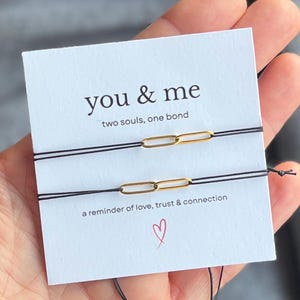 Könnte beinhalten: Zwei schwarze Armbänder mit goldfarbenen Kettenglied-Anhängern werden auf einer weißen Karte präsentiert. Auf der Karte steht "you & me" mit dem Text "two souls, one bond" und "a reminder of love, trust & connection". Auf der Karte ist ein kleines rotes Herz abgebildet.
