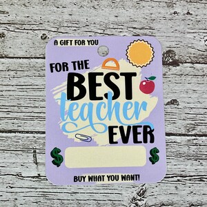 K&ouml;nnte beinhalten: Eine lilafarbene Geschenkkarte mit dem Text "A Gift For You" oben. Die Karte sagt "For The Best Teacher Ever" in schwarz-wei&szlig;er Schrift auf blau-gelbem Hintergrund. Auf beiden Seiten eines leeren Feldes zum Eintragen eines Betrags befinden sich Dollarzeichen. Der Text "Buy What You Want!" befindet sich am unteren Rand der Karte.
